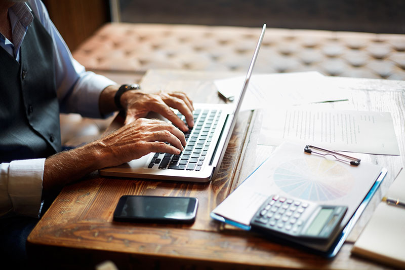 A man types on his laptop at a table covered in work papers